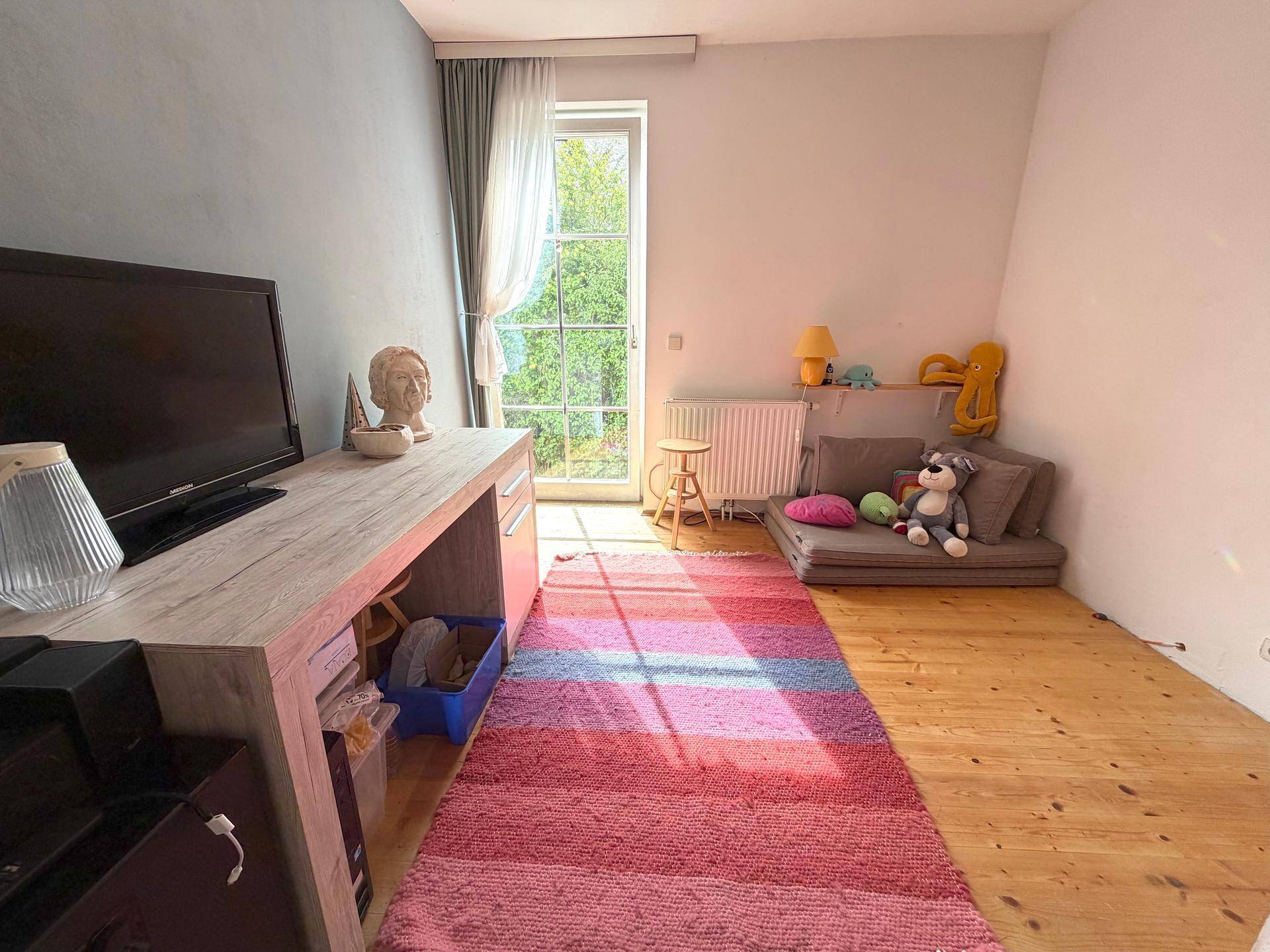 Helles Kinderzimmer mit Holzboden, Schreibtisch und Zugang zum Balkon.