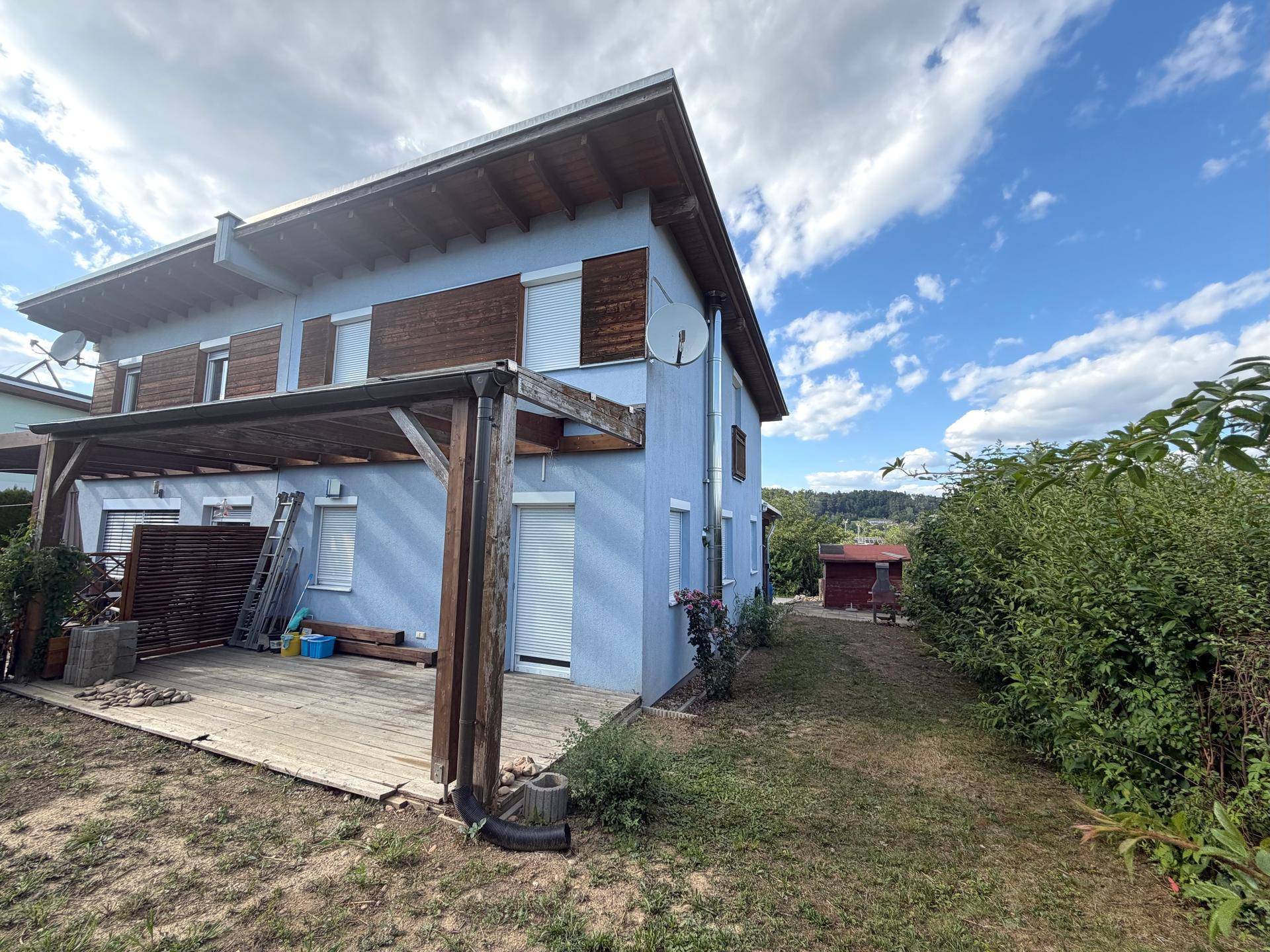 Seitenansicht des Hauses mit Garten, Schuppen und Holzterrasse unter blauem Himmel.
