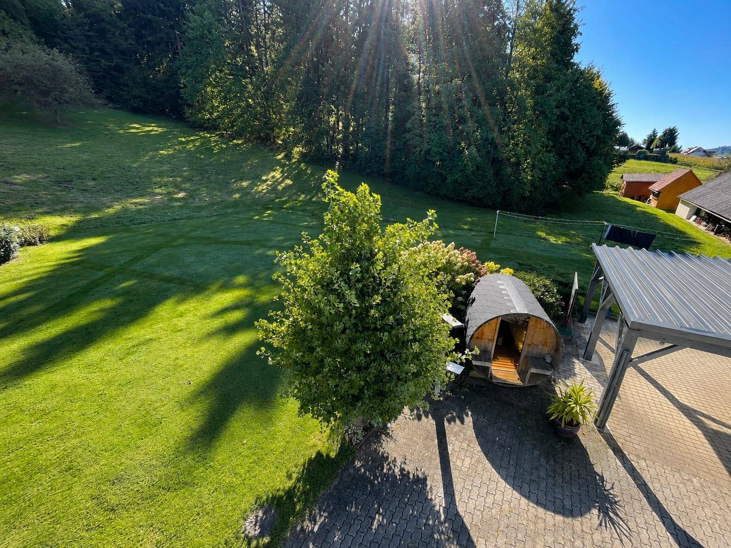 Weitläufiger Garten mit Rasenfläche, Bäumen, einer Fasssauna und einem gepflasterten Bereich.