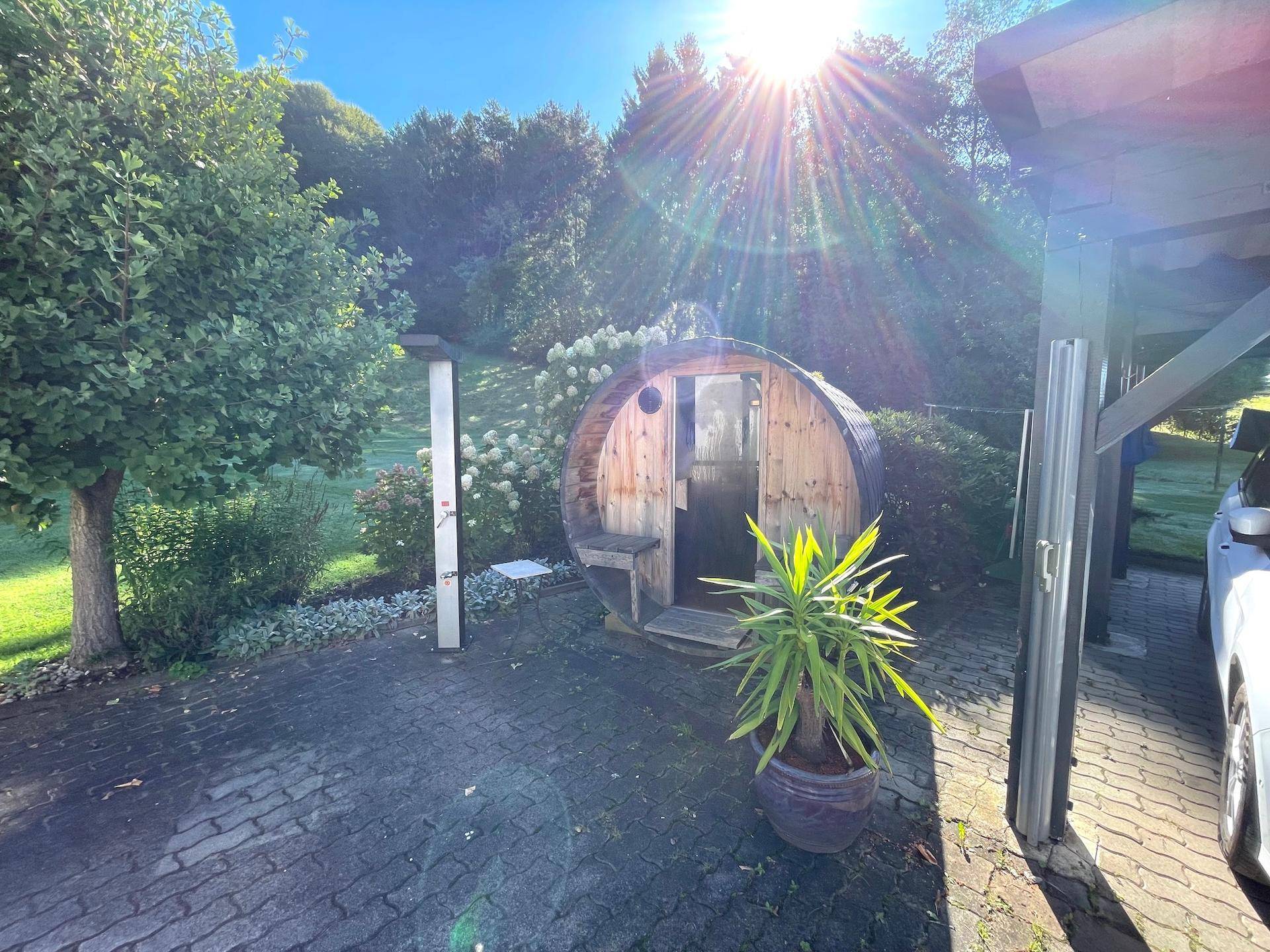 Gepflasterter Bereich mit einer Fasssauna, Topfpflanzen und Blick auf den grünen Garten.