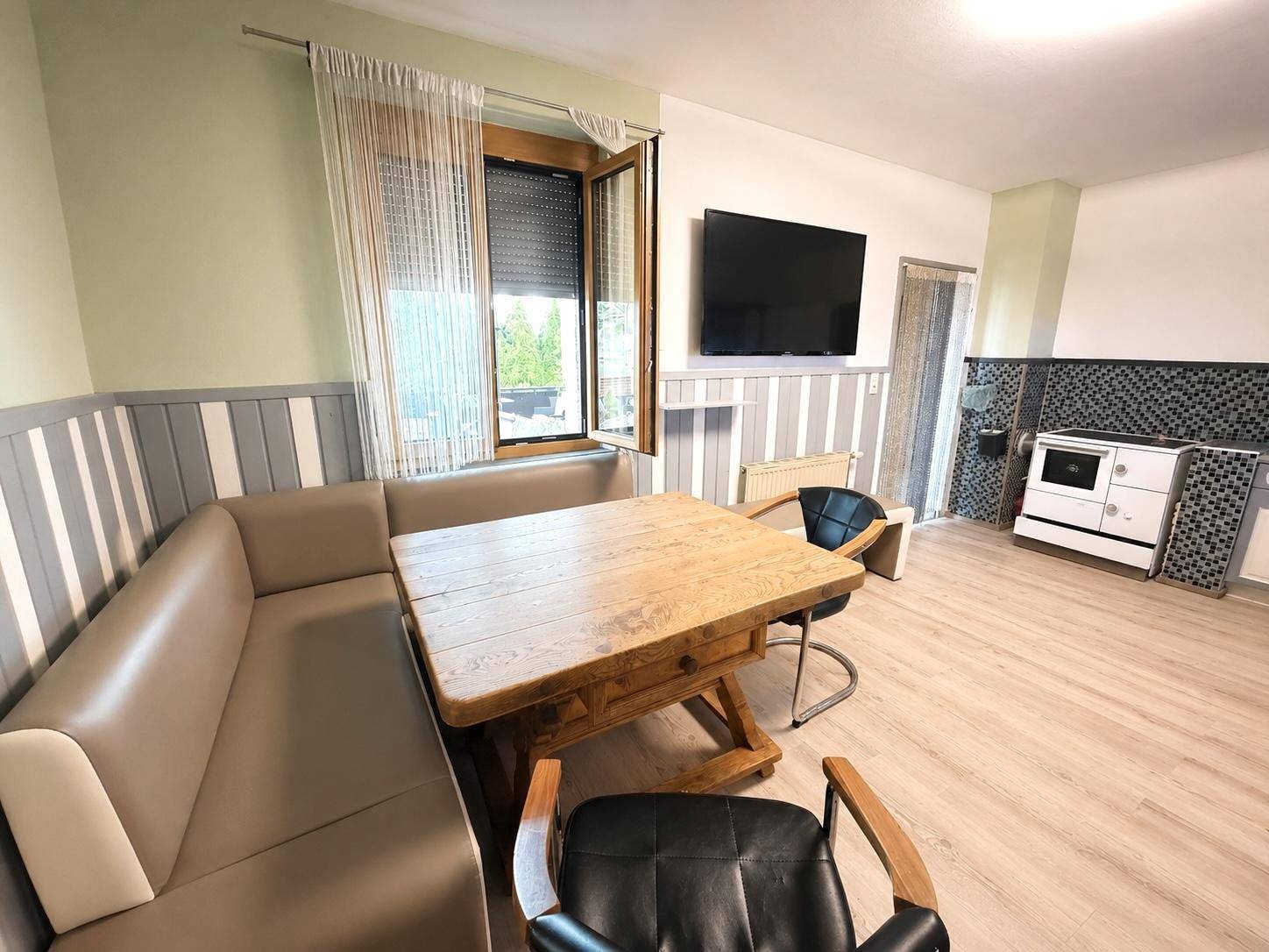 Gemütlicher Essbereich mit Holztisch, Eckbank und Fernseher, daneben ein traditioneller Ofen.