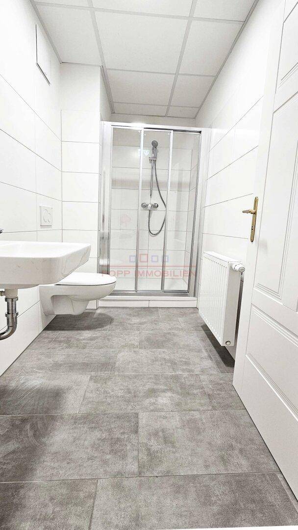 Modernes Badezimmer mit grauen Bodenfliesen, weißen Wandfliesen, wandhängendem WC und einer Duschkabine.