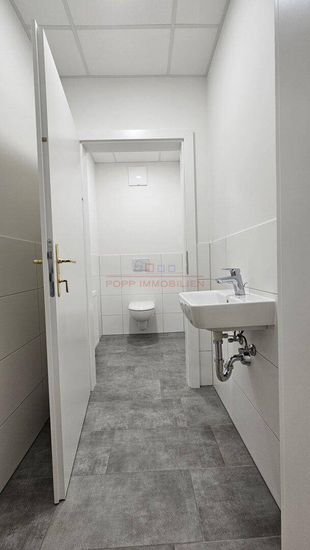 Heller Sanitärbereich mit grauen Bodenfliesen, Waschbecken und separatem WC-Raum, der durch eine Tür zugänglich ist.