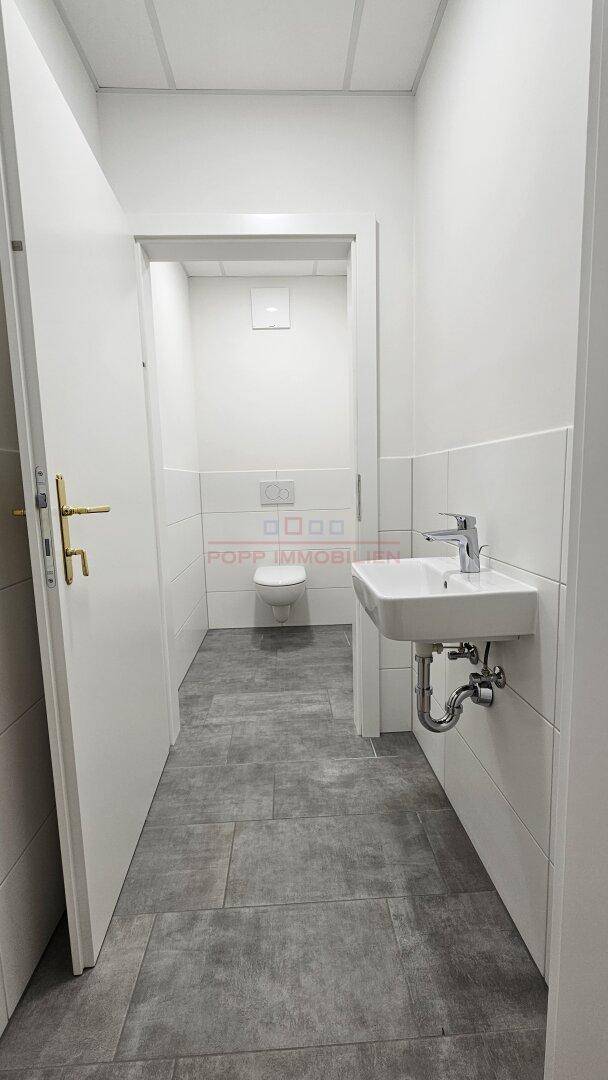 Sauberer Sanitärbereich mit grauen Bodenfliesen und einem Waschbecken, der zu einem separaten WC-Raum führt.