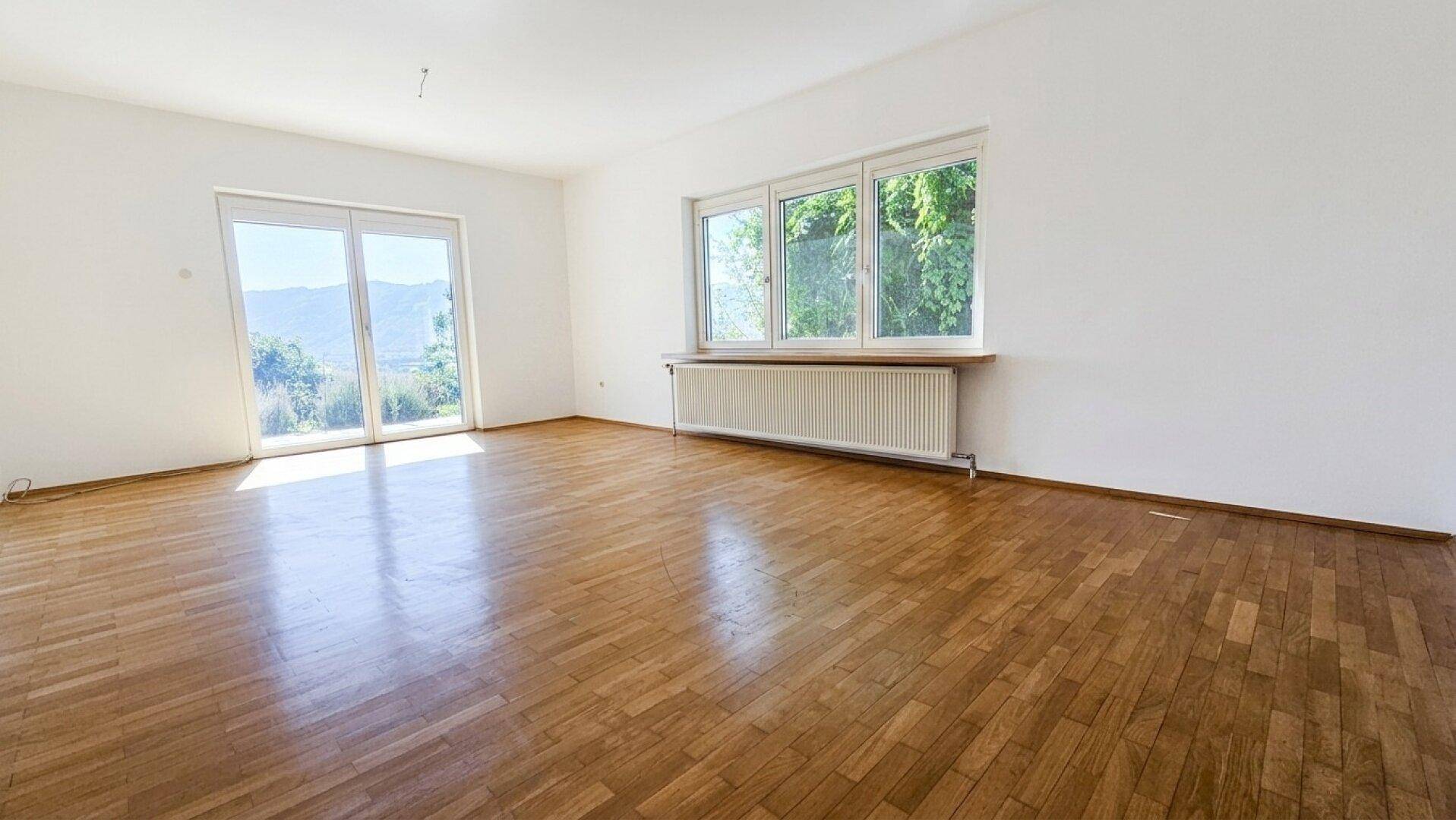 Helles, leeres Wohnzimmer mit Parkettboden, großen Fenstern und Zugang zum Außenbereich.