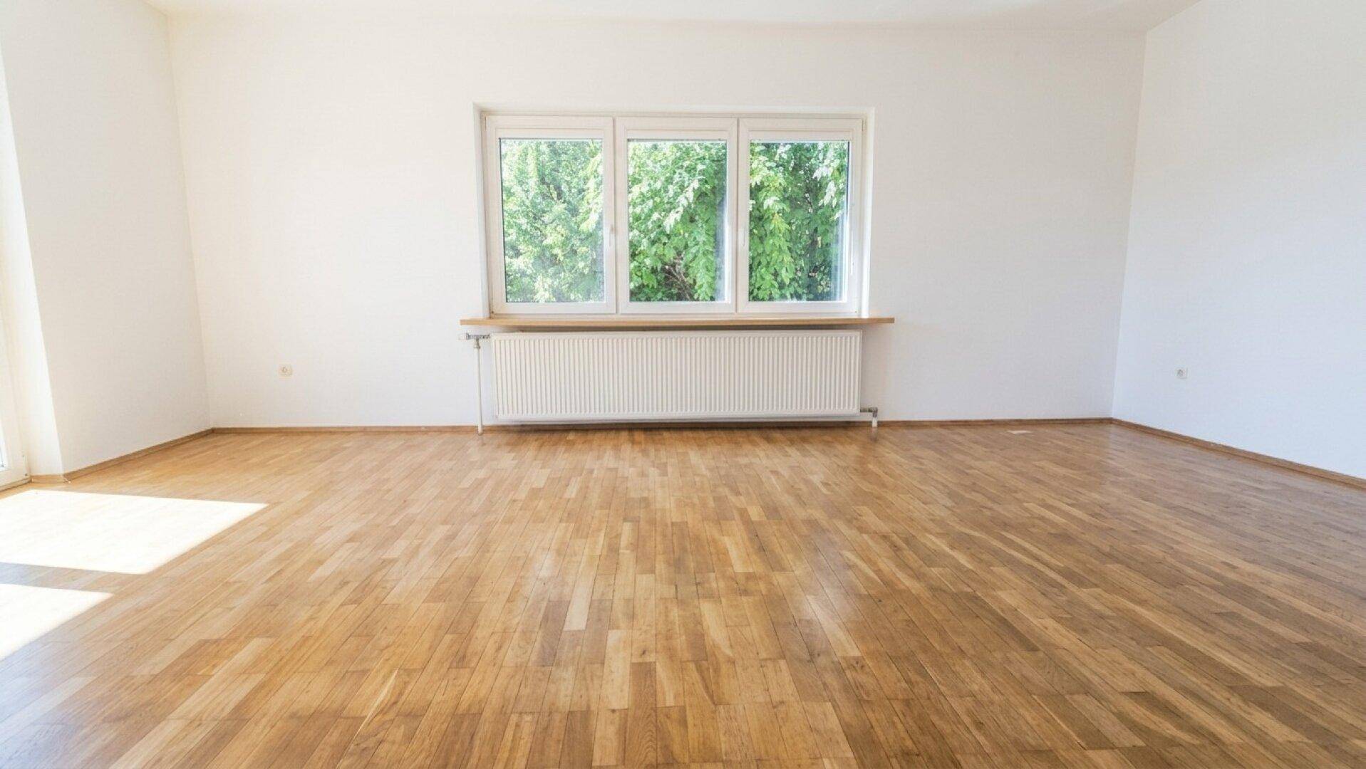 Geräumiges Zimmer mit hellem Parkettboden und großem Fenster mit Blick ins Grüne.