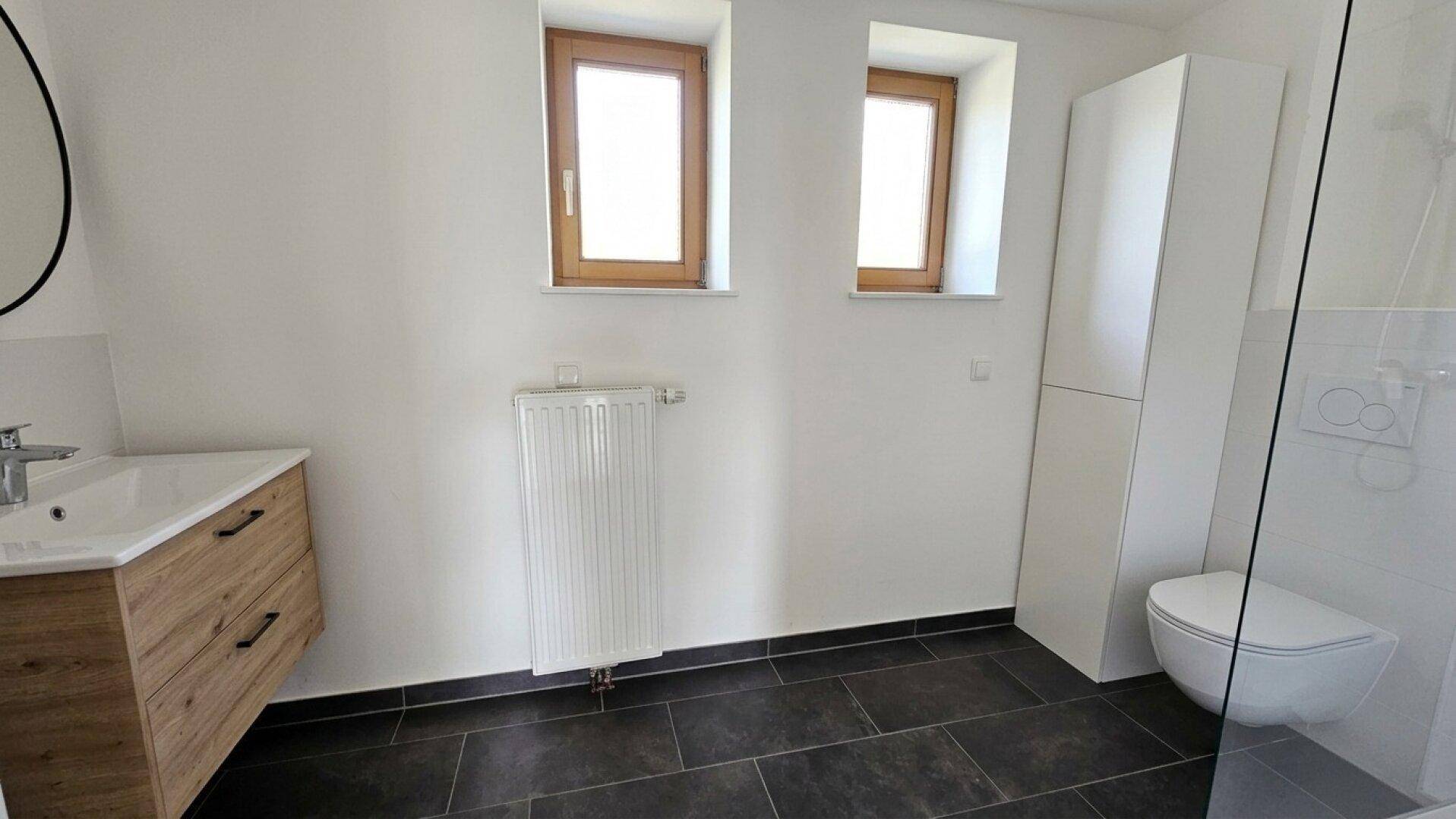 Modernes Badezimmer mit dunklen Fliesen, zwei Fenstern, Waschtisch und Duschbereich.