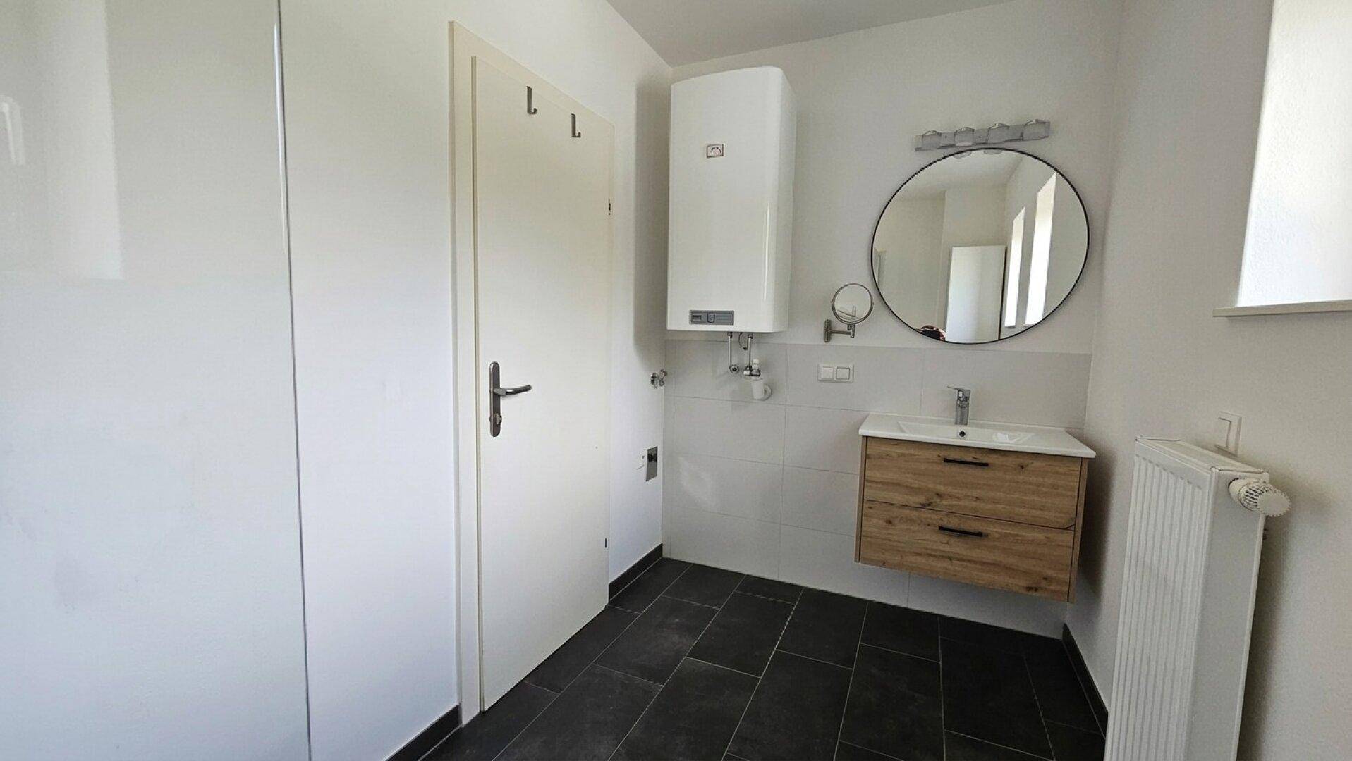 Modernes Badezimmer mit dunklem Fliesenboden, Waschtisch, rundem Spiegel und Warmwasserbereiter.