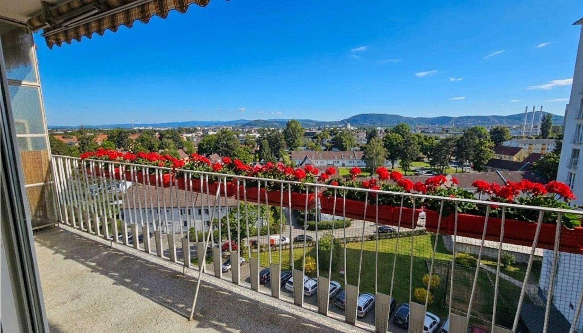 Sonniger Balkon mit Blumenkästen und weitem Blick über die Stadt und grüne Hügel.