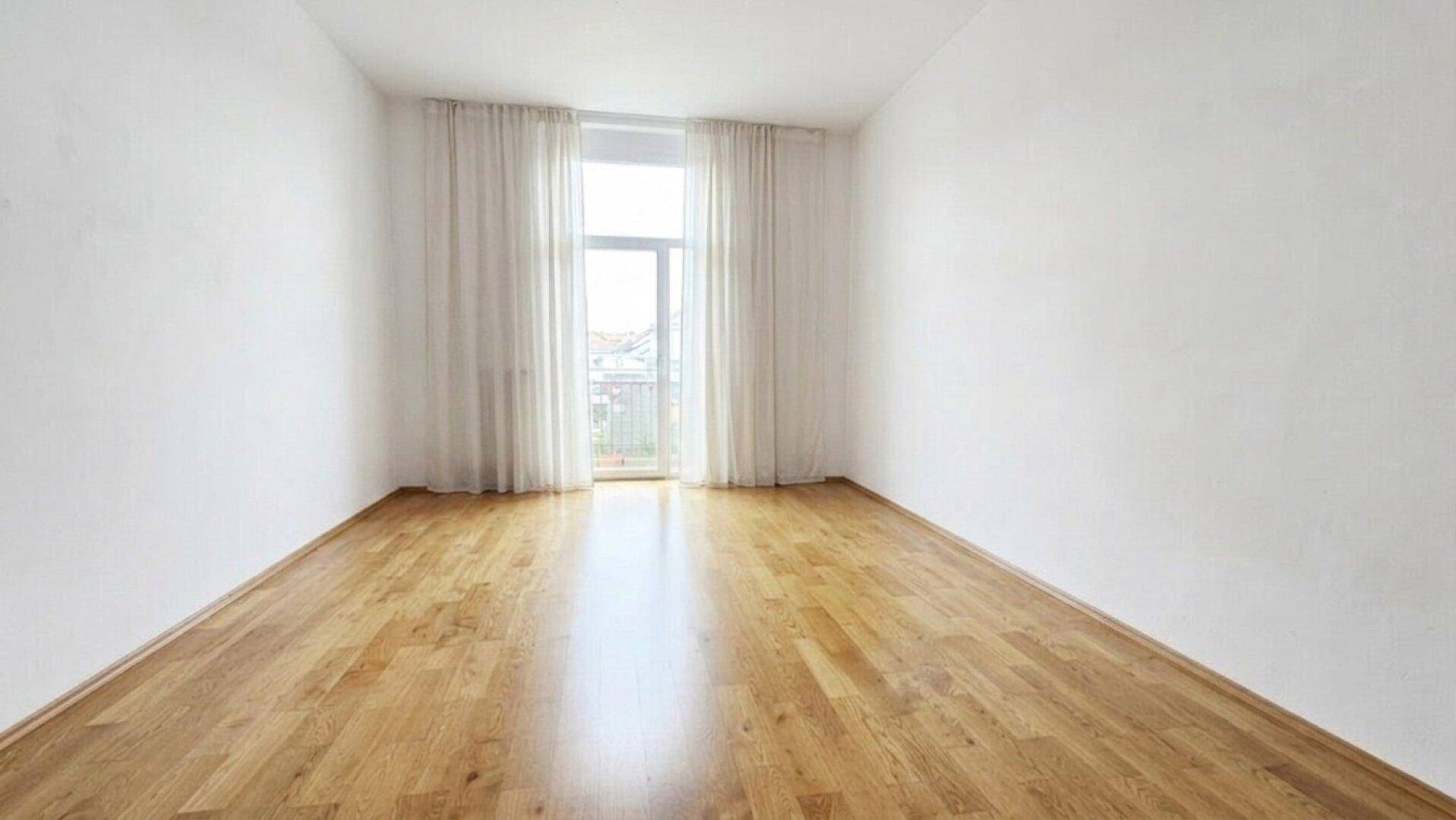 Leeres, helles Zimmer mit Holzboden und großem Fenster, das viel Licht hereinlässt.