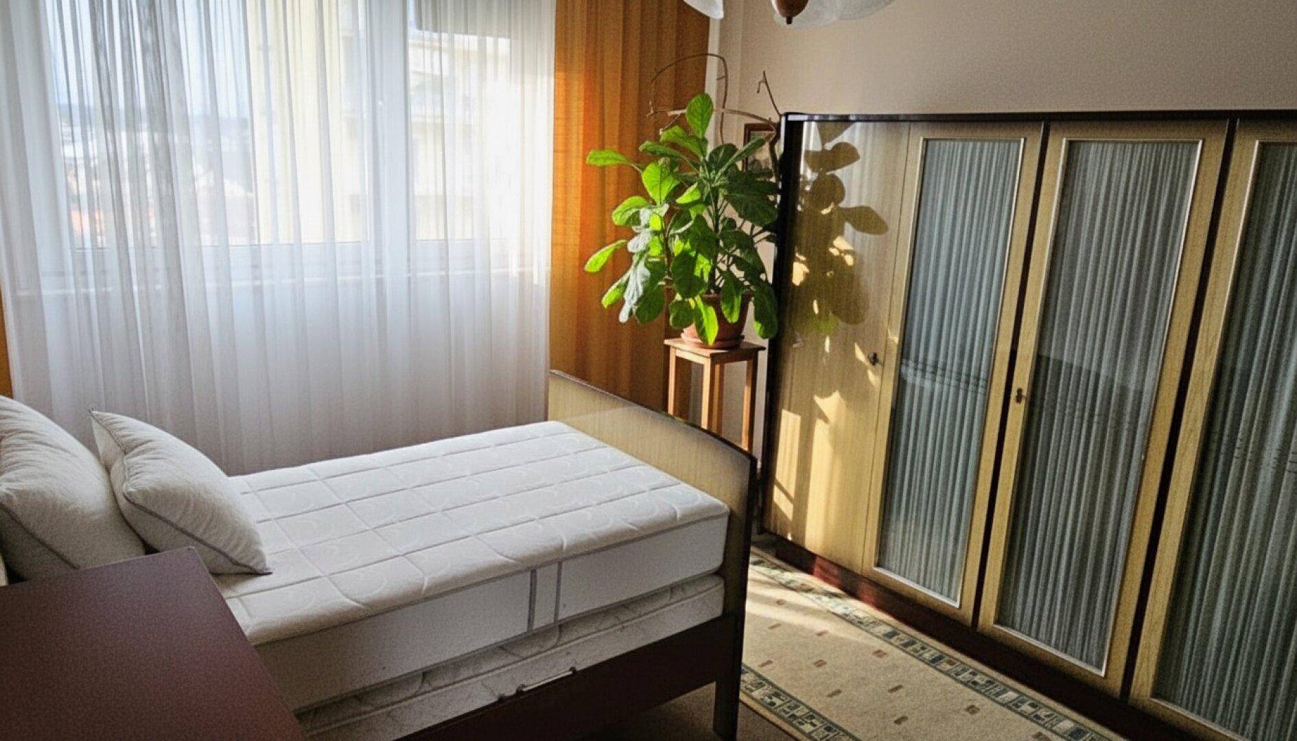Helles Schlafzimmer mit Einzelbett, Kleiderschrank und großen Fenstern mit Vorhängen.