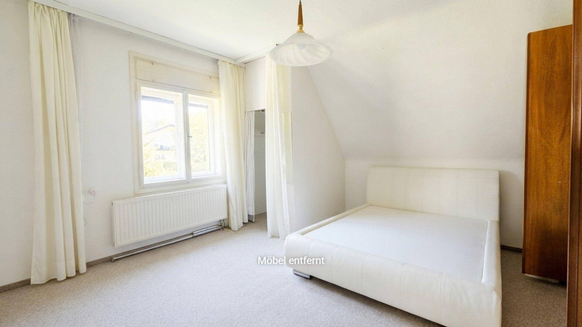 Schlafzimmer mit Teppichboden, weißem Bett und Dachschräge, hell durch großes Fenster.
