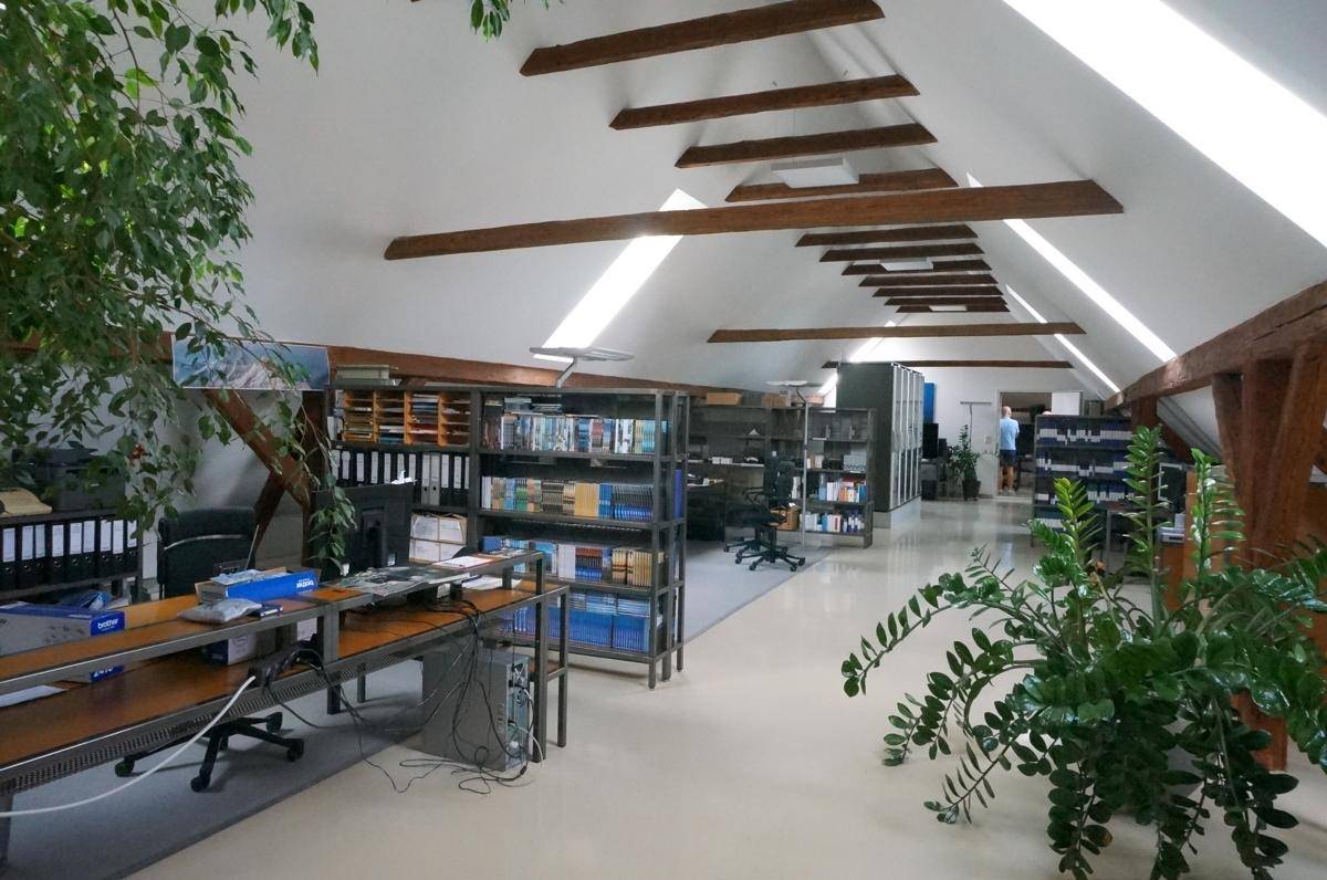 Langer Bürobereich im Dachgeschoss mit Regalen, Arbeitsplätzen und sichtbaren Holzbalken.