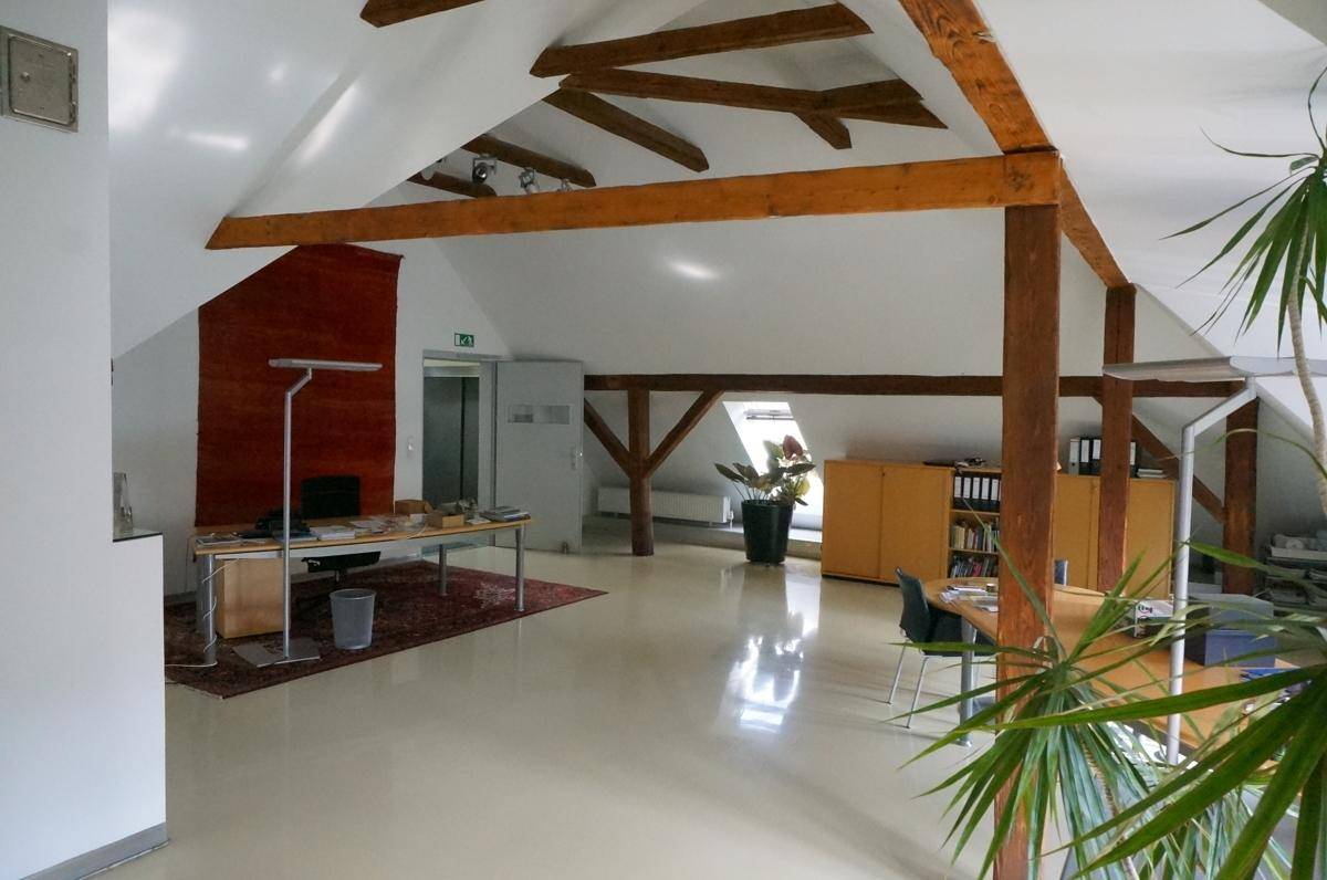 Dachgeschossbüro mit sichtbaren Holzbalken, Teppich und einem Aufzug im Hintergrund.