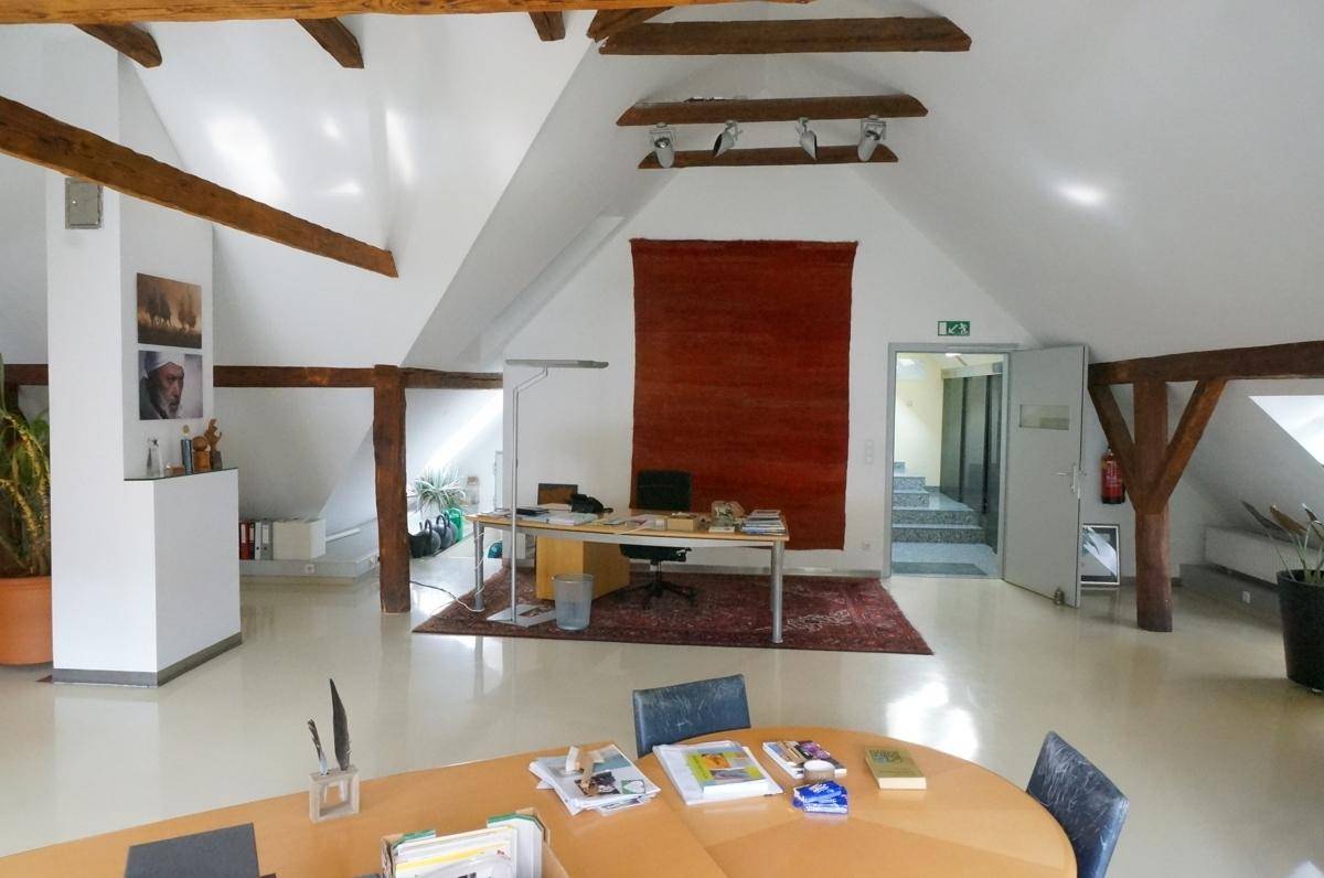 Geräumiges Dachgeschossbüro mit Holzbalken, Teppich und Zugang zum Treppenhaus.