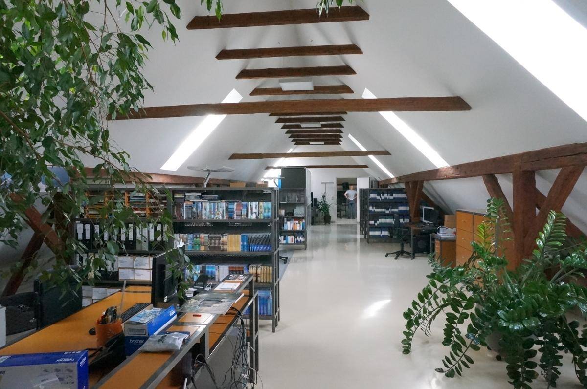 Langer Bürobereich im Dachgeschoss mit Regalen, Arbeitsplätzen und sichtbaren Holzbalken.