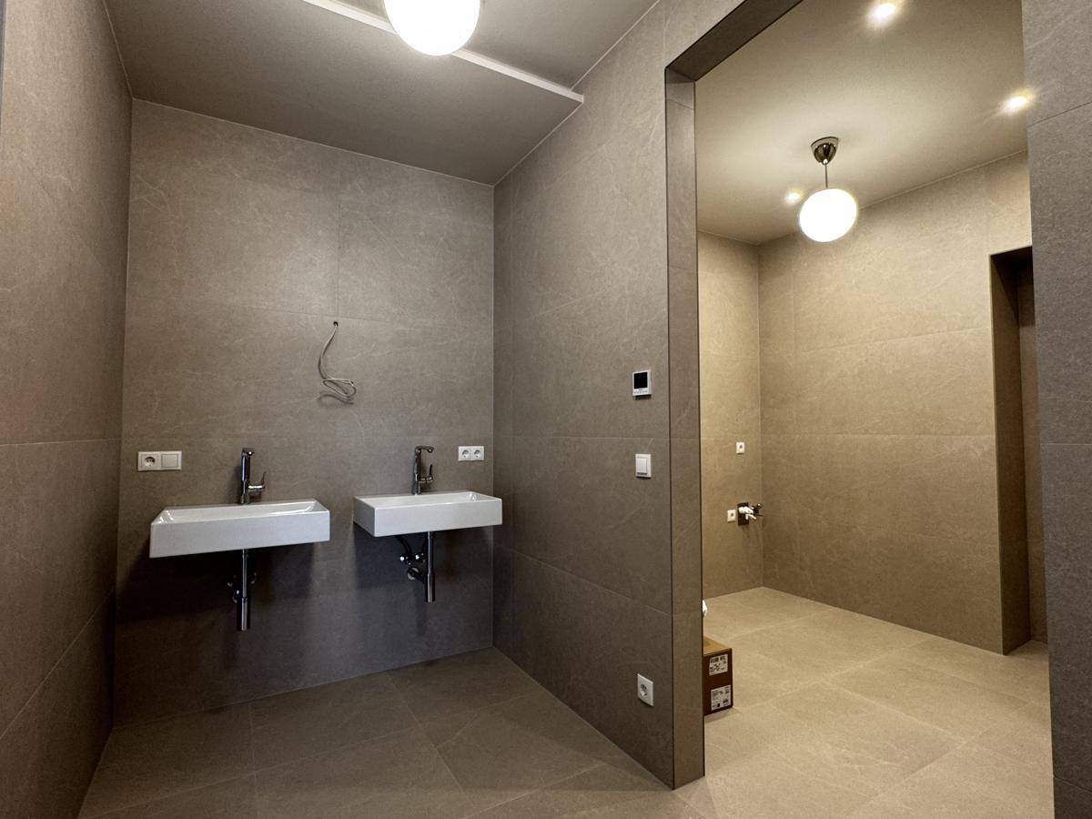 Geräumiges Badezimmer mit zwei modernen Waschbecken und durchgängiger grauer Fliesenverkleidung.
