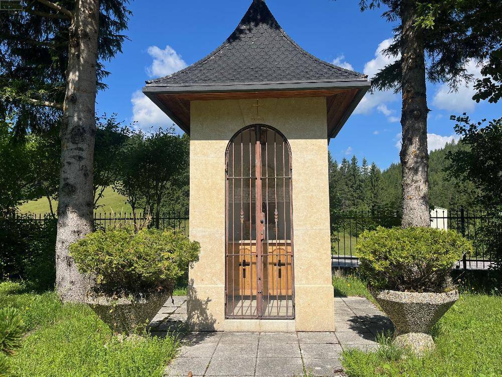 Kleine Kapelle oder Schrein mit schmiedeeisernem Gitter und zwei Bäumen davor in ländlicher Umgebung.
