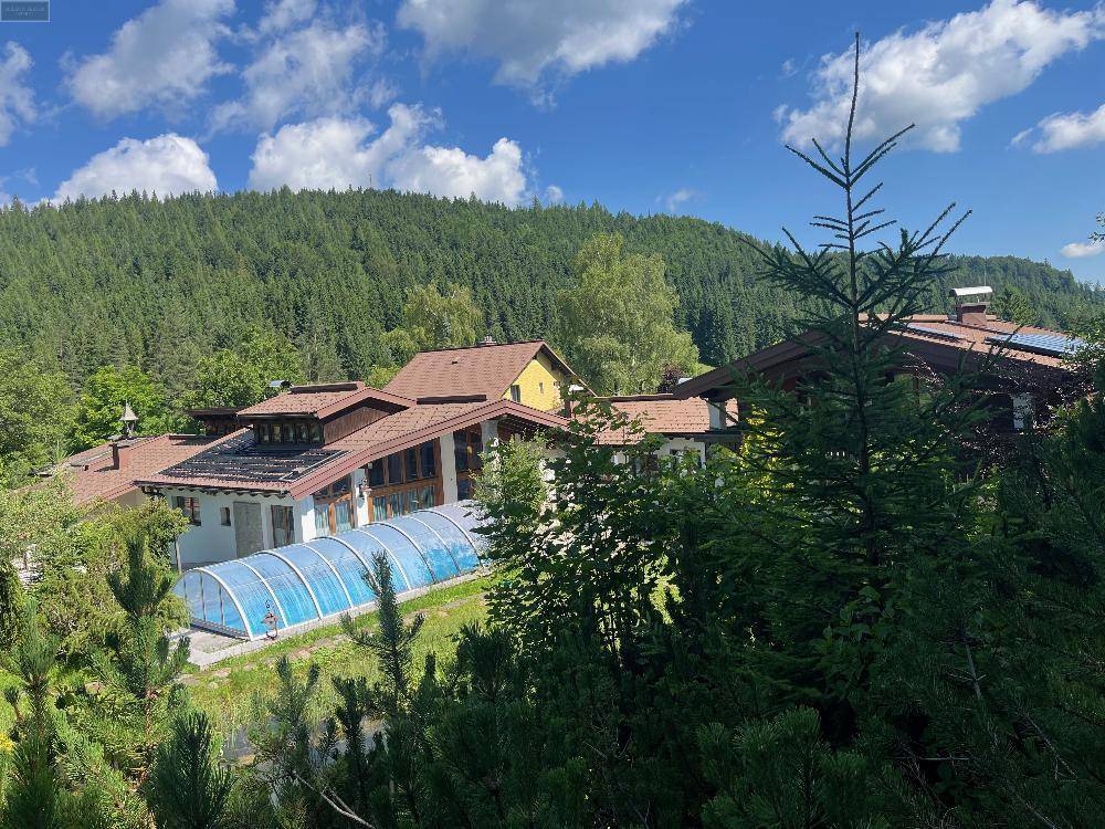 Luftaufnahme des Anwesens mit seinen Dächern und dem umgebenden dichten Wald unter blauem Himmel.