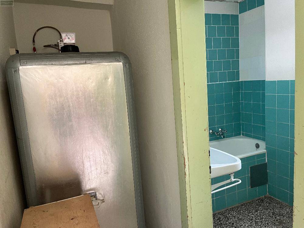 Abstellraum mit Warmwasserspeicher und Blick auf ein Badezimmer mit türkisfarbenen Fliesen und Badewanne.