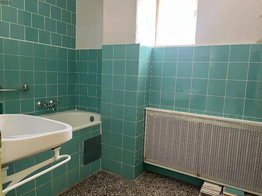 Badezimmer mit türkisfarbenen Wandfliesen, Badewanne, Waschbecken und Heizkörper, in renovierungsbedürftigem Zustand.