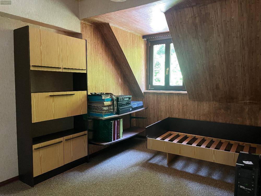 Dachzimmer mit schräger Decke, Holzverkleidung und einem einfachen Bettgestell, renovierungsbedürftig.