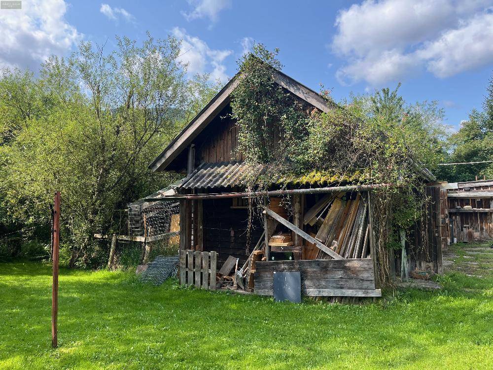 Alte, überwucherte Holzscheune mit Vordach und gelagertem Material, in ländlicher Umgebung.