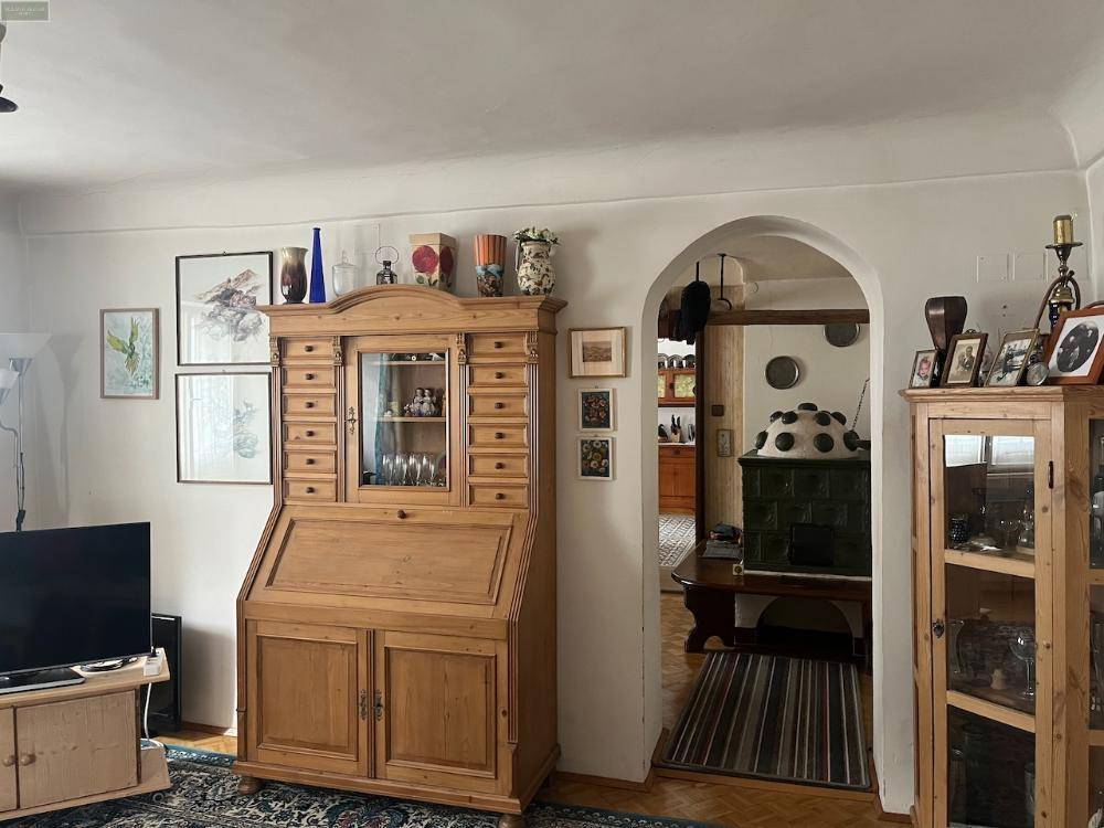 Wohnzimmer mit großem Holzsekretär und Fernsehmöbel, ein Bogen führt zu einem Raum mit Kachelofen.