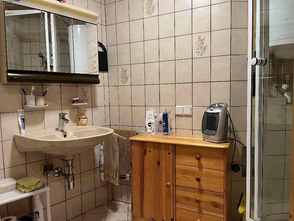 Badezimmer mit beigefarbenen Fliesen, Waschbecken, Spiegelschrank und Duschkabine, ein kleiner Heizlüfter steht bereit.