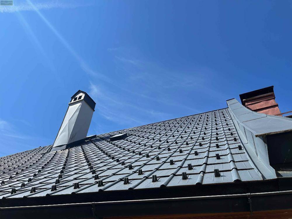 Dach eines Hauses mit grauen Ziegeln und zwei Schornsteinen unter klarem blauem Himmel.