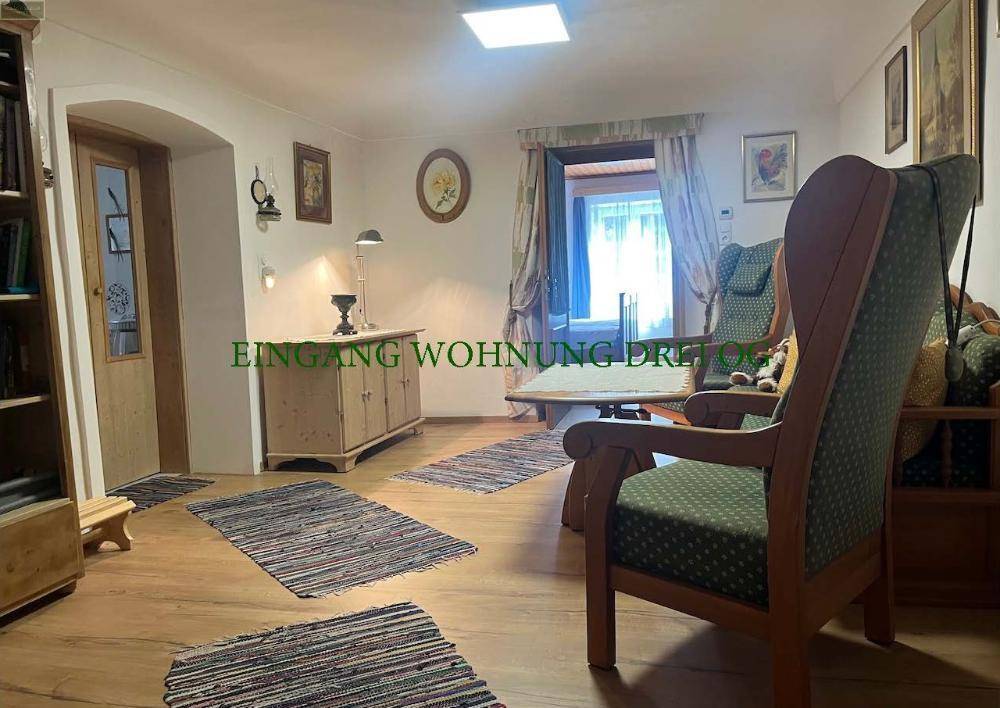 Wohnzimmer mit Holzboden, quadratischer Deckenleuchte und einem Sessel, ein Eingangsbereich ist sichtbar.