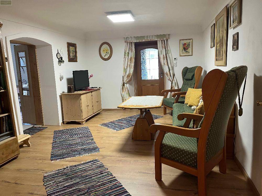 Wohnzimmer mit Holzboden, Fernseher auf einem Holzschrank und mehreren Sesseln, eine Tür führt nach draußen.