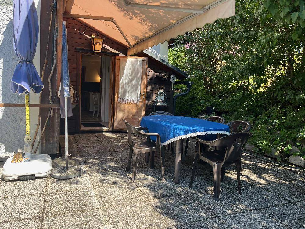 Außenterrasse mit gepflastertem Boden, einem Tisch mit Stühlen und Sonnenschirm, umgeben von viel Grün.