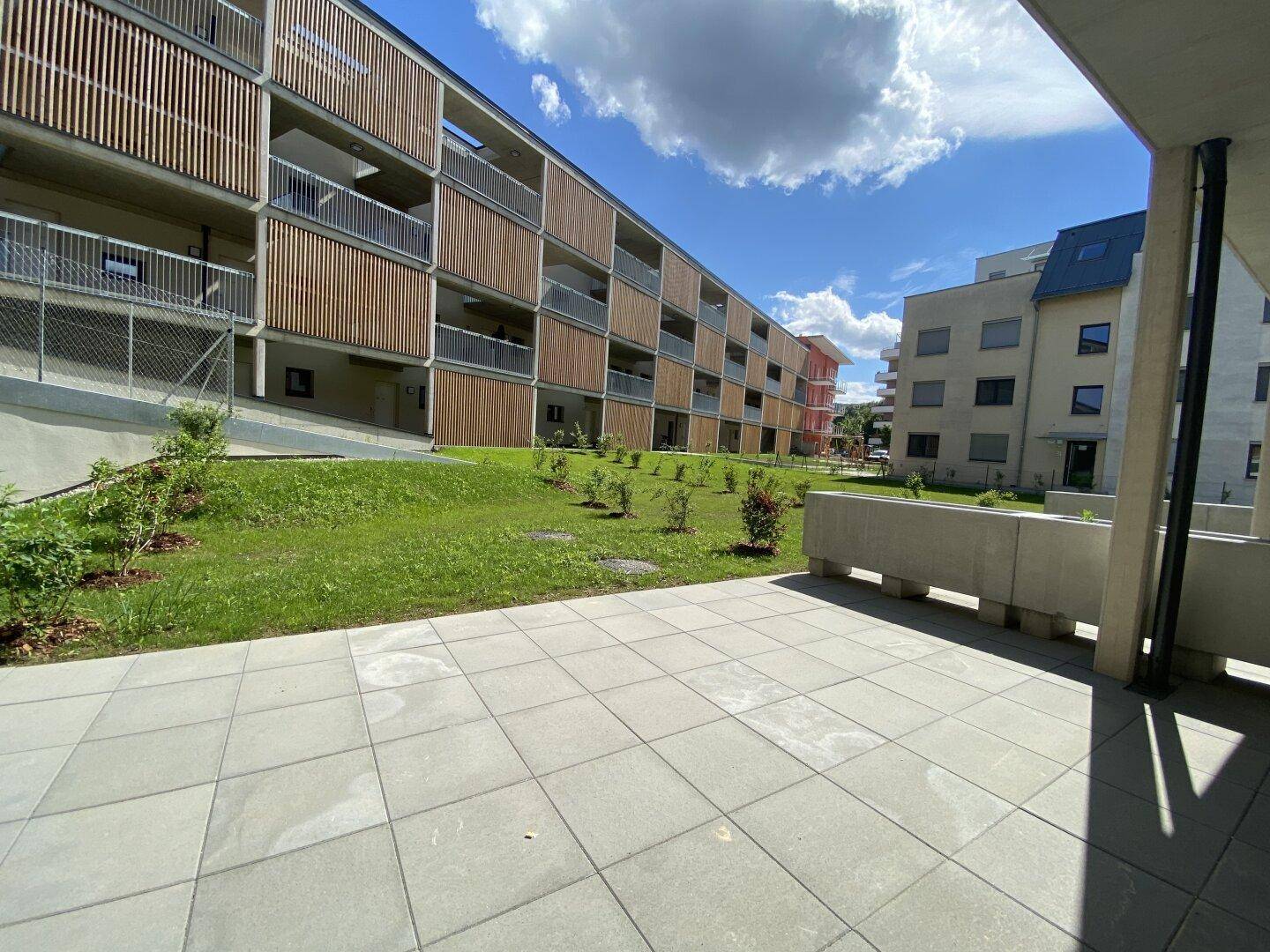 Geräumige Terrasse mit Blick auf die grüne Rasenfläche und moderne Wohngebäude unter blauem Himmel.