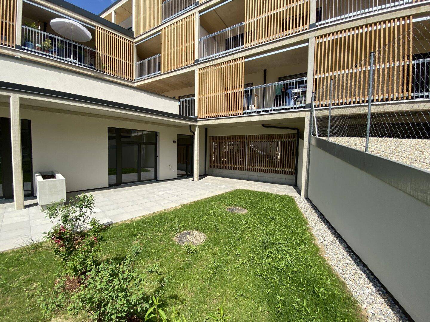 Außenbereich mit Rasenfläche und gepflasterter Terrasse vor einem modernen Apartmentgebäude.