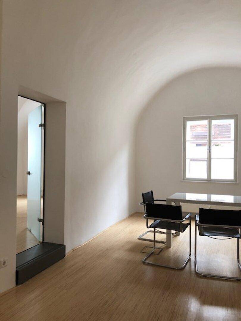 Geräumiger Büroraum mit Gewölbedecke, Holzboden und einer modernen Glastür, die zu einem weiteren Bereich führt.