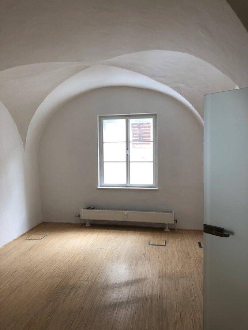 Büroraum mit charakteristischer Gewölbedecke, Holzboden und einem Fenster, unter dem ein Heizkörper installiert ist.