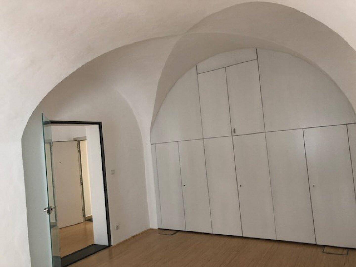 Raum mit eleganter Gewölbedecke, Holzboden und einer Wand mit integrierten, weißen Einbauschränken für Stauraum.