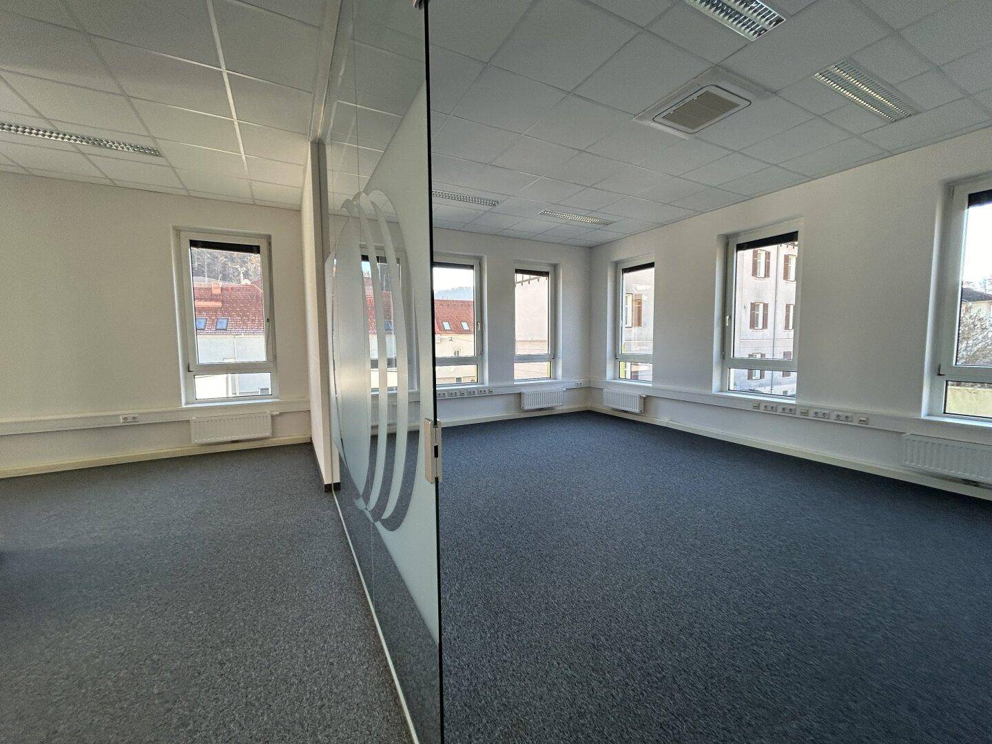 Helles Büro mit grauem Teppichboden, großen Fenstern und einer modernen Glaswand, die den Raum unterteilt.