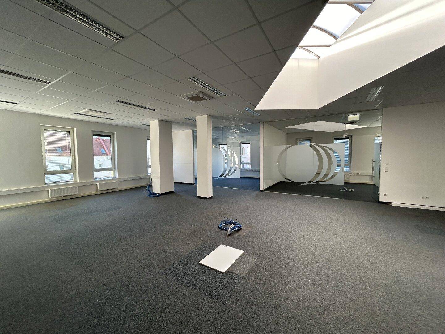 Großes, offenes Büro mit Teppichboden, Säulen, Glaswänden und einem Oberlicht, das viel Tageslicht hereinlässt.