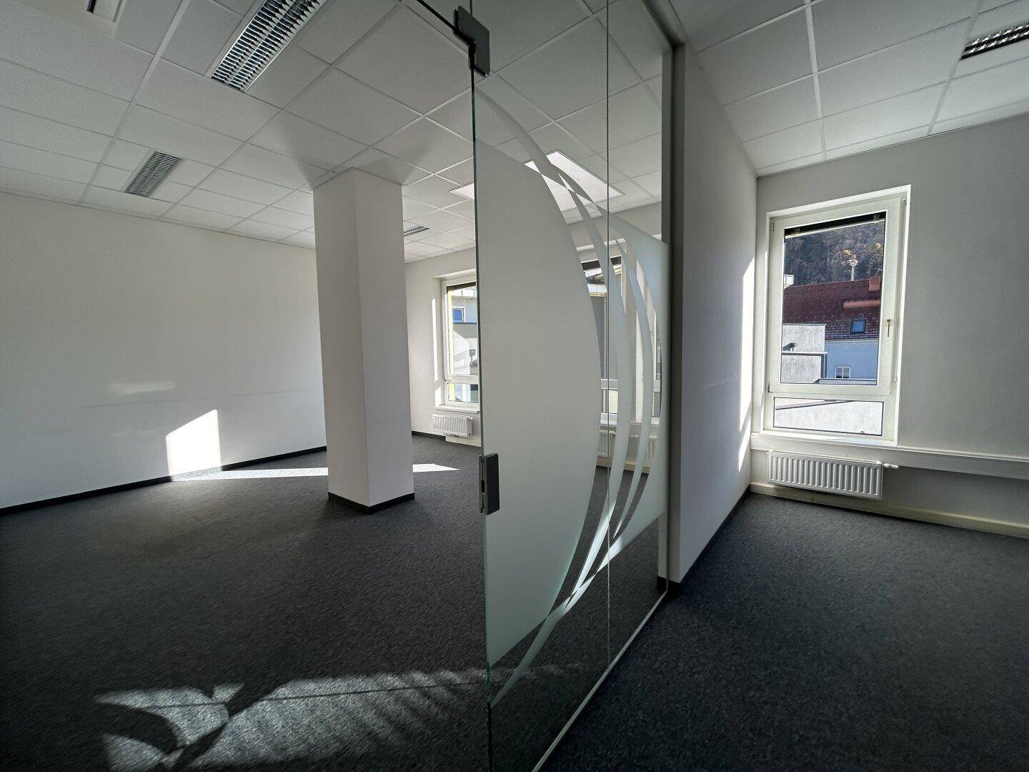 Büroraum mit grauem Teppichboden, einer Säule und einer Glastür, die den Zugang zu einem weiteren Bereich ermöglicht.