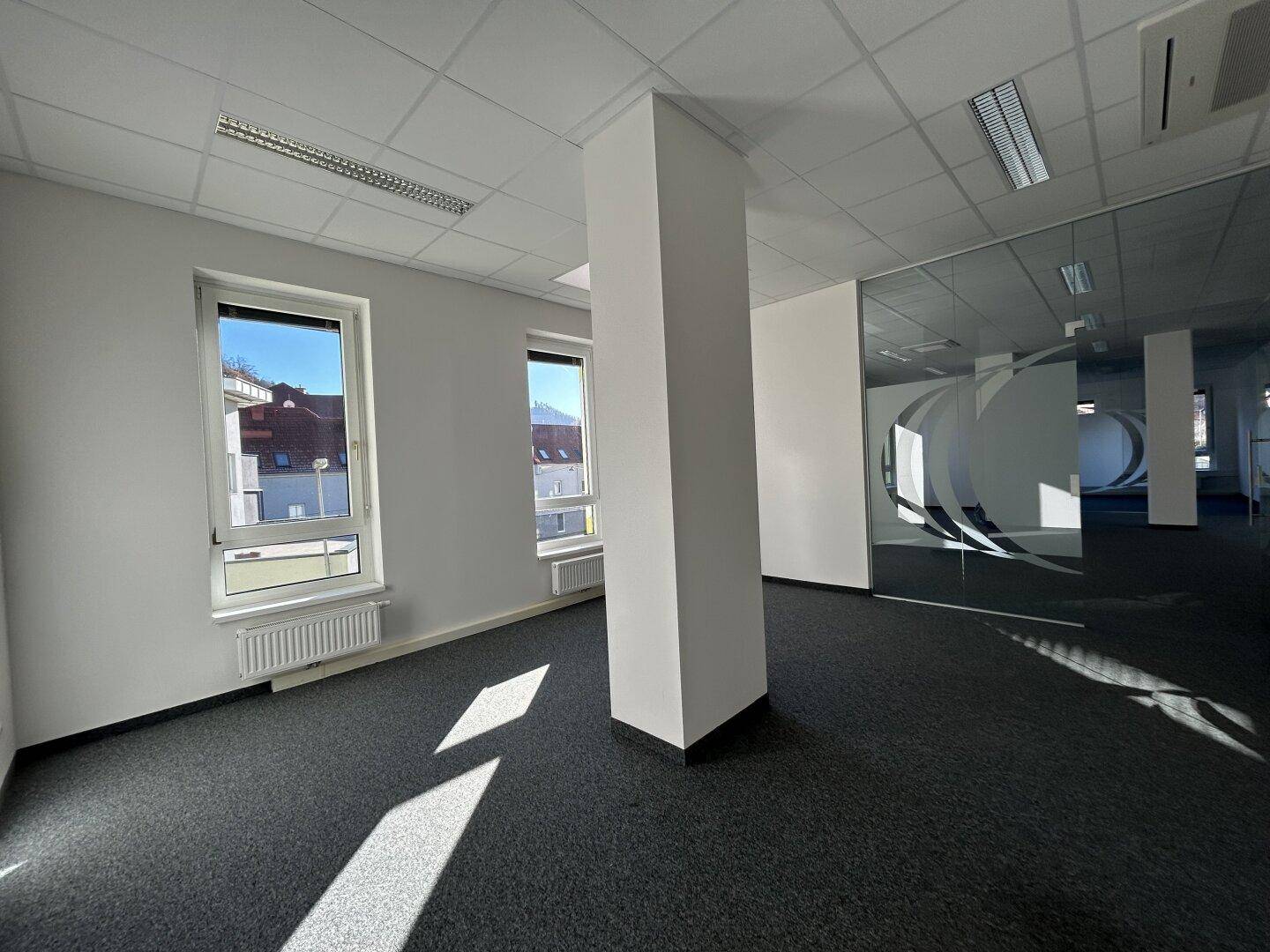 Modernes Büro mit grauem Teppichboden, einer tragenden Säule und eleganten Glaswänden, die den Raum strukturieren.