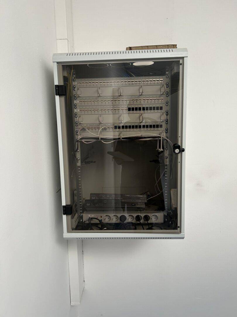 Technischer Raum mit einem an der Wand montierten Serverschrank, der Netzwerkgeräte und Verkabelung enthält.