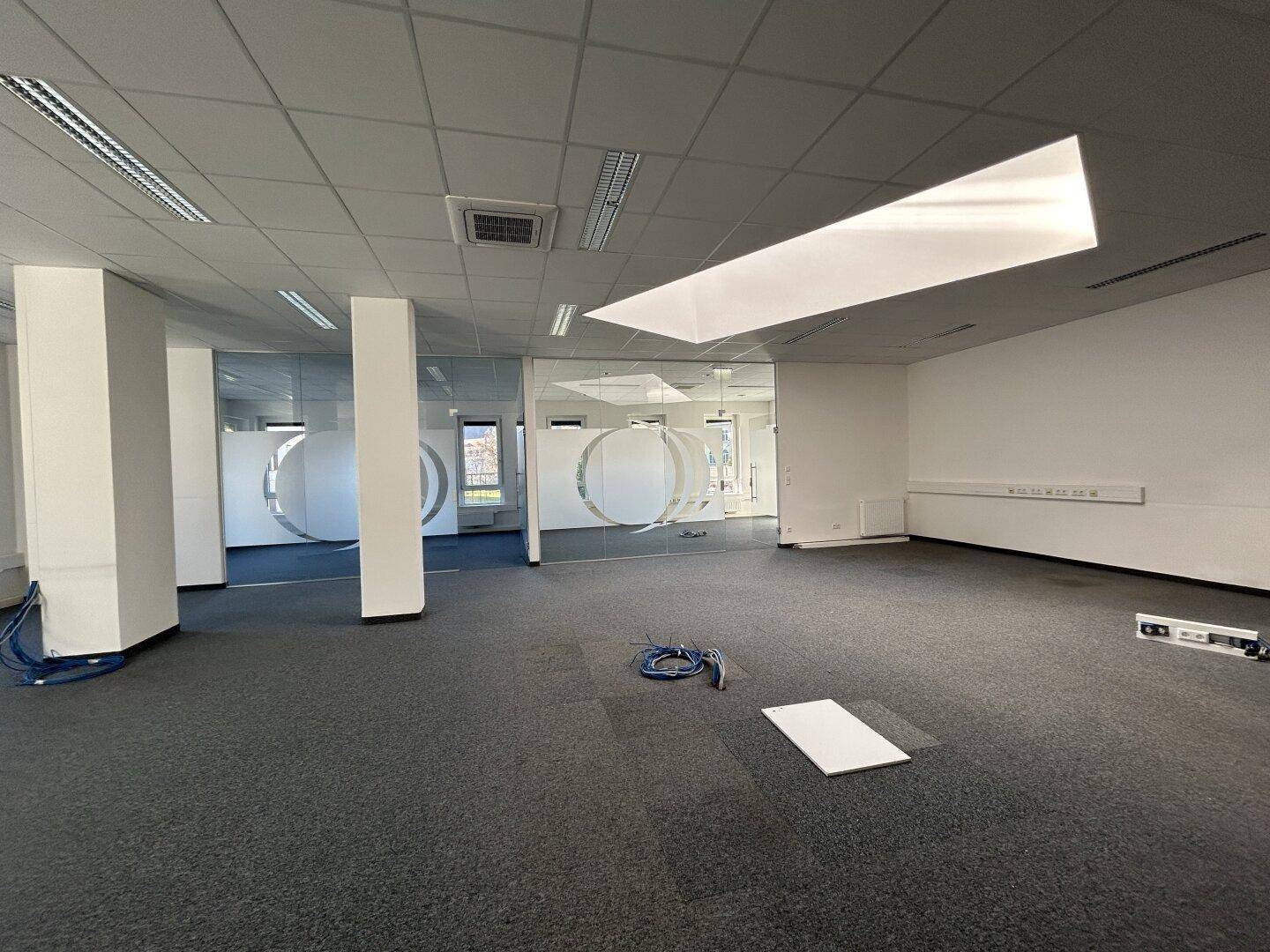 Großes Büro mit Teppichboden, Säulen und Glaswänden, das eine moderne und flexible Arbeitsumgebung bietet.