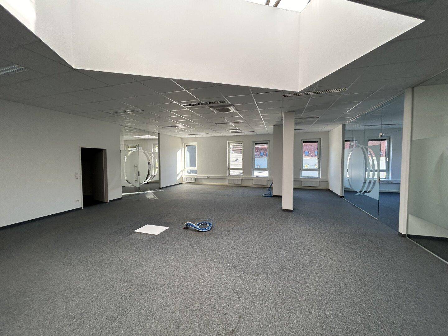 Weitläufiges, offenes Büro mit Teppichboden, Glaswänden und einem großen Oberlicht, das für eine helle Arbeitsumgebung sorgt.
