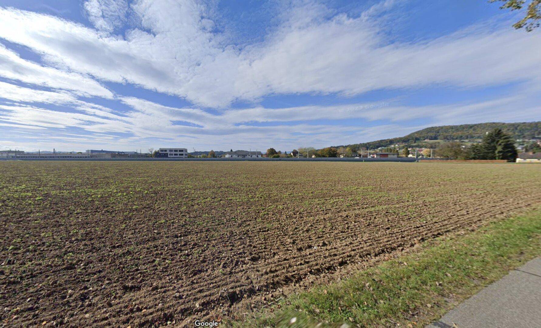 Weites Feld mit Blick auf die umliegende Landschaft und bewaldete Hügel unter blauem Himmel.