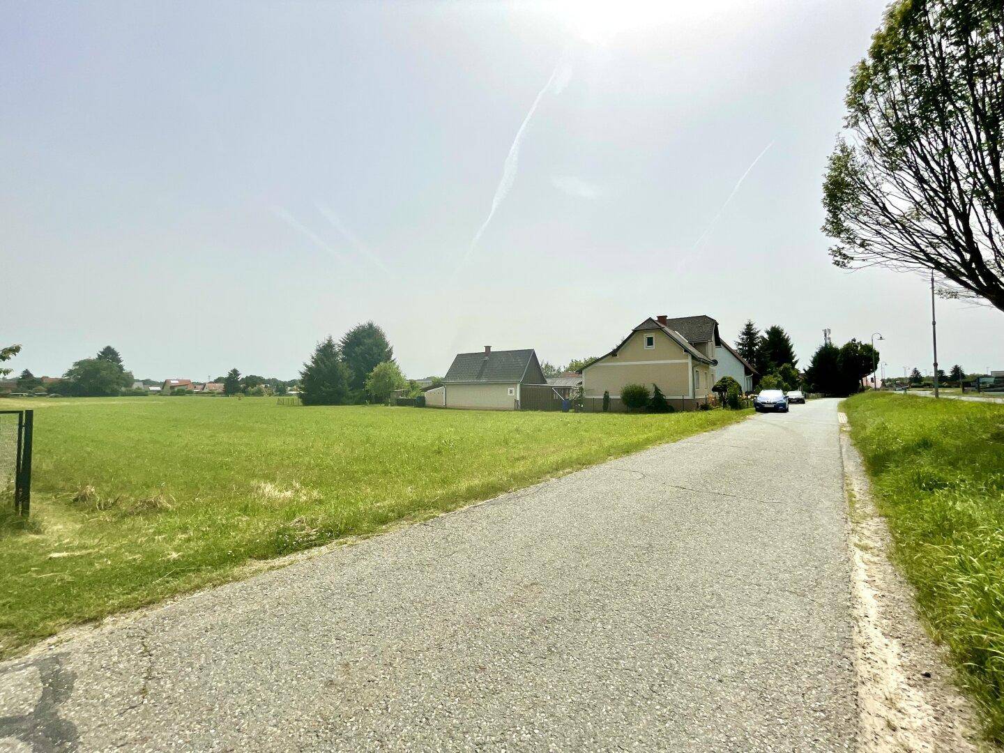 Weitläufiges grünes Feld mit Häusern im Hintergrund und einer ländlichen Straße.