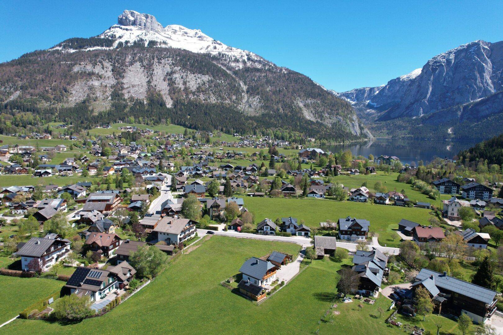 Luftaufnahme einer malerischen Siedlung am Seeufer, umgeben von grünen Hügeln und schneebedeckten Bergen.