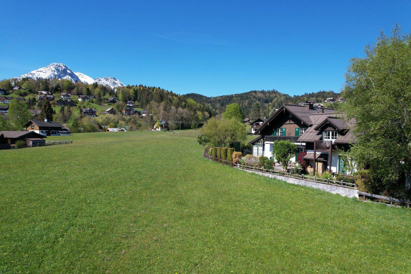 Weitläufige grüne Wiesen mit traditionellen Häusern am Waldrand und Bergen im Hintergrund unter blauem Himmel.