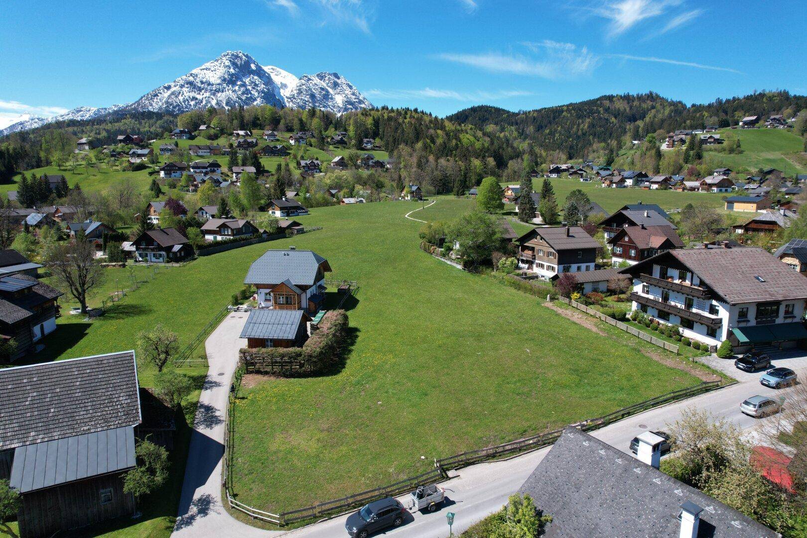 Dorfansicht aus der Vogelperspektive mit verstreuten Häusern, grünen Feldern und einer Straße vor Bergkulisse.